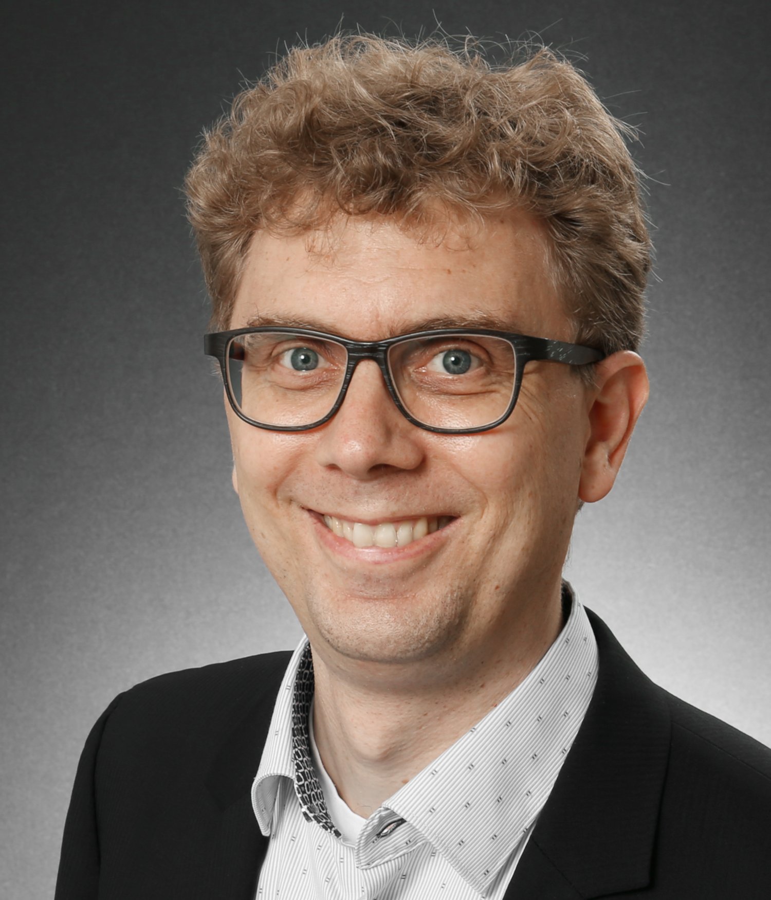 Smiling man with curly light brown hair and glasses wearing a black blazer and a white patterned shirt against a grey gradient background